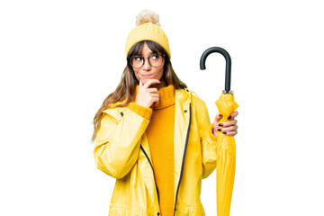 Young caucasian woman with rainproof coat and umbrella over isolated chroma key background having doubts and thinking - obrazy, fototapety, plakaty