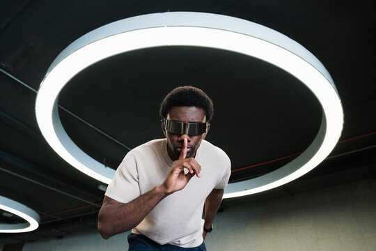 Young man with futuristic cyber glasses under modern ring lamp putting forefinger on lips