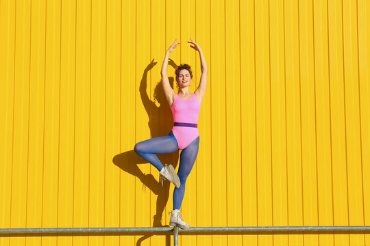Young woman wearing bodysuit and practicing ballet on railing in front of yellow wall