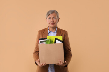Unemployed senior woman with box of personal office belongings on beige background