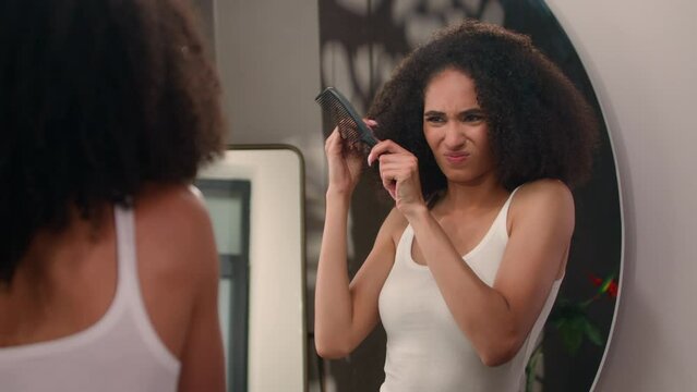 Frustrated exhausted mad angry sad African American woman trying to comb curls suffer with haircare problem looking at mirror reflection girl female suffering try brush curly tangled hair in bathroom