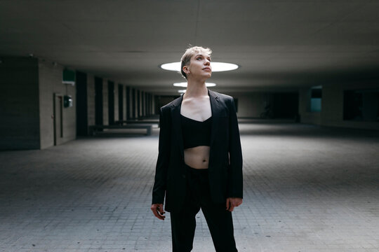 Thoughtful trans young man wearing black suit standing on floor in basement