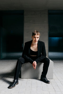 Confident trans young man wearing black suit sitting on seat in basement