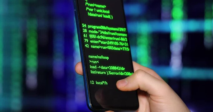 Close up of cell phone in the hand of a young hacker or programmer. On the background screen and phone screen program code and software. Hacking or programming concept