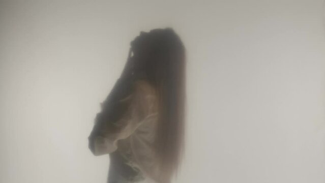 Side view of silhouette of woman unbuttoning buttons on blouse and gently running her hand over her neck. A woman in steam or fog behind a transparent frosted glass or curtain close up. HDR BT2020 HLG