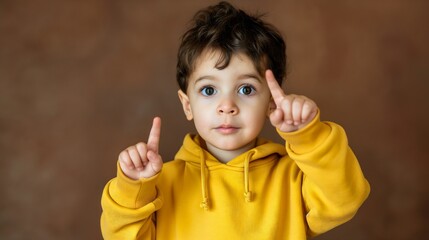 Cute young boy wearing a yellow hoodie pointing up with his finger. Smiling and looking at the camera. Studio shot, wall in the background. Hand gesture, here, showing direction, recommended advice