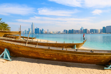 Traditional Arabic boats docked on a sandy beach at Marina Mall Island, with the waterfront corniche and skyline across the sea in Abu Dhabi, United Arab Emirates.