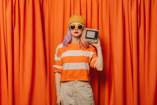 Woman wearing knit hat holding retro style television set in front of orange curtain
