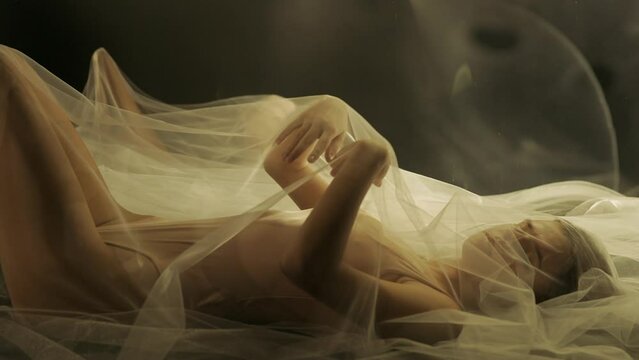 Side view of a young woman on a bed, under a transparent air cloth. A woman illuminated by light is reflected in a liquid mirror. The woman gently touches the white fabric with her hands. Slow motion.