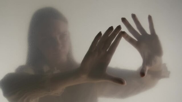 Silhouette of ghost woman in fog behind frosted curtain or glass. Woman's hands touching the glass in close up. Concept of afterlife, otherworld, ghosts. HDR BT2020 HLG Material.
