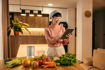 An asian woman is preparing healthy smoothie at home while smiling at tablet and scrolling on it.