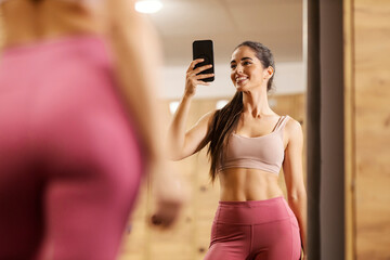 Portrait of a sportswoman taking selfies in a mirror in a gym fitting room.