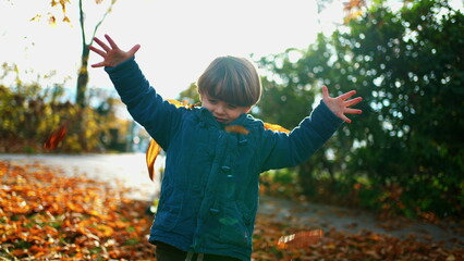 Playful child putting orange leaves in head while standing out sunlit park during autumn fall...