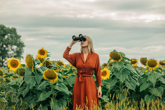 Woman in red dress looking through binoculars at sunflower field
