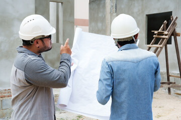 Two-man home design engineers standing in front actual work, looking at plans talking about construction that is about be completed, wear safety helmets protect their heads while in construction site.