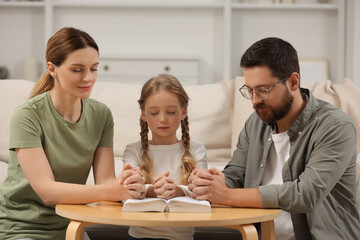 Girl and her godparents praying over Bible together at table indoors