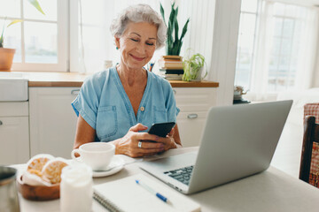Cheerful elderly lady with gray hair in denim shirt sitting at table in front of laptop at home,...