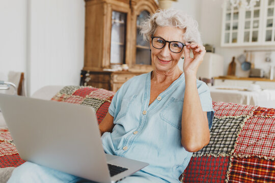 Indoor portrait of adorable lady of 60s using laptop, reading news, e-books, watching tutorials, browsing social media,holding laptop on laps, touching her glasses looking at camera