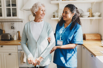 Medical assistance at home. Pretty african american female volunteer in blue uniform helping senior caucasian woman walk with walking stick, holding her by hand, looking at each other with smile - obrazy, fototapety, plakaty