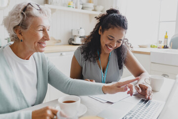 Side view of two people sitting at kitchen table in front of laptop, african american female social worker volunteer helping senior lady filling in form, elderly woman pointing at screen with smile - 723146100