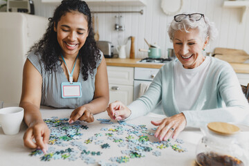 Portrait of african american female social worker volunteer playing puzzle game with cheerful senior lady, having fun, laughing searching for right piece sitting together at kitchen table
