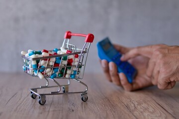 A shopping cart overflowing with colorful pills on a wooden table and hand using calculator. Rising...