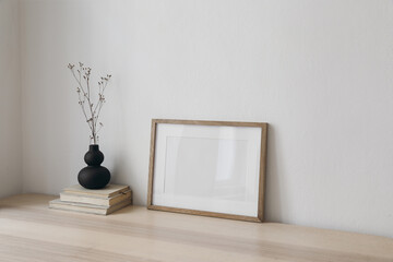 Blank horizontal wooden picture frame mockup. Organic shaped black vase with dry grass on table,...