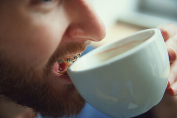 Cropped portrait of bearded man with dental braces drinking hot coffee or tea from white porcelain...