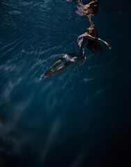 Underwater shoot of beautiful ballerina in white dress swimming and dancing in water through sunbeams.
