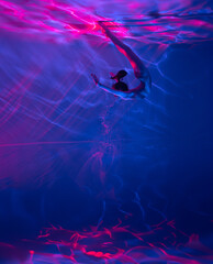 Underwater shoot of ballerina swimming and dancing in water through pink and red light rays.