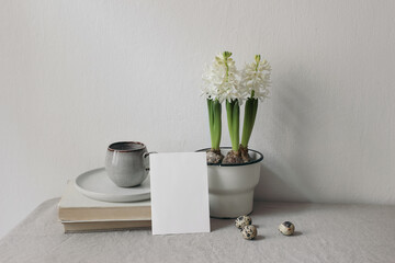 Easter spring still life. Blank greeting card, invitation mockup. Cup of coffee, books on linen table cloth. White hyacinth in flower pot. Quail eggs. Home office concept, Scandinavian interior.