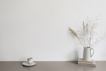 Autumn breakfast still life scene. Ceramic jug with dry grass and old books. Cup of coffee on beige table. Blank white wall mockup. Scandinavian interior, boho home. No people. Neutral design