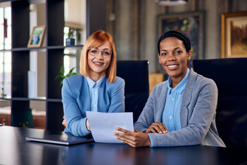 A portrait of two business females, having a meeting, holding one document, and smiling at the camera.