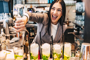 Woman barman preparing alcoholic cocktail and pouring it into glass with crushed ice, mint and lime...