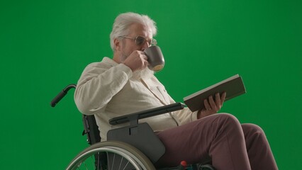 Portrait of disabled man isolated on chroma key green screen close up. Senior man sitting in wheelchair holding book drinking tea from mug.