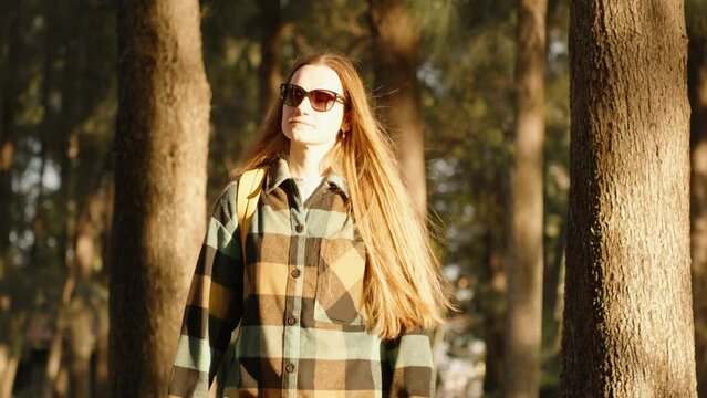 A young woman with sunglasses walks among the trees in the forest during a vibrant sunset.