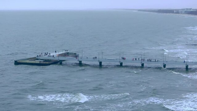 In a January drone film, the Baltic Sea coastline unfolds with Kolobrzeg pier in the distance. Strolling people, flying seagulls, golden sands, and stormy waves paint a winter seascape.