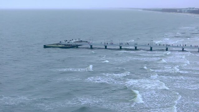 In a January drone film, the Baltic Sea coastline unfolds with Kolobrzeg pier in the distance. Strolling people, flying seagulls, golden sands, and stormy waves paint a winter seascape.