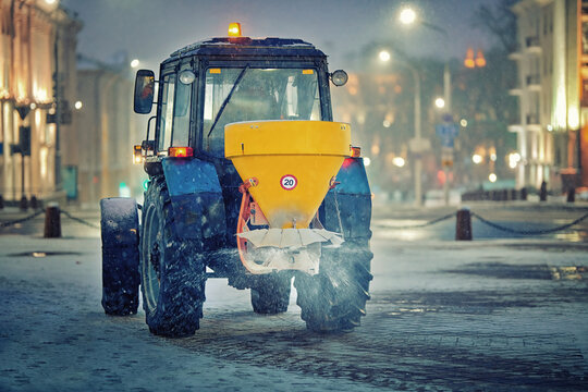 Tractor deicing street, spread salt. Salt spreader machine work at night, salting paving slabs, tactor spreading rock salt on sidewalk, winter road maintenance, prevent slipping. Selective focus