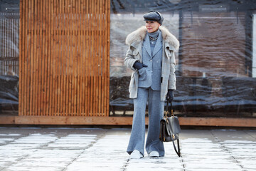 Stylish mature woman stands full length on street in winter, wears fashionable multilayer outfit,...