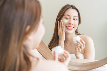 Facial beauty skin care, smile of pretty asian young woman in bathrobe looking at mirror, hand applying moisturizer lotion on her face, holding jar of skin cream before makeup cosmetic routine at home