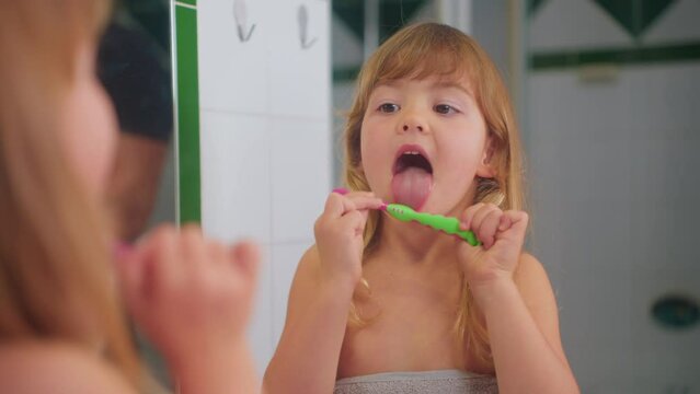 Cute little girl sitting in front of the mirror in the bathroom in a towel cleans her tongue with a toothbrush