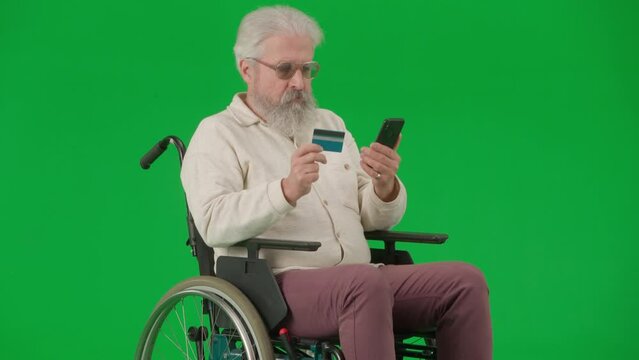 Portrait of aged man on chroma key green screen. Senior man in wheelchair typing bank card number in smartphone, buying online.