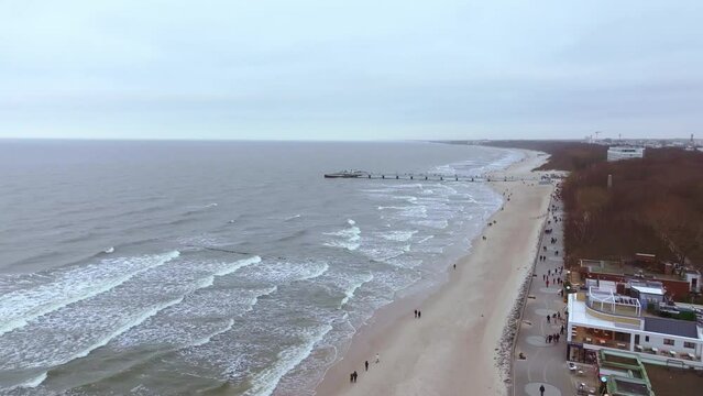 A January drone film captures Baltic Sea coastline with distant Kolobrzeg pier. People stroll on the beach amid flying seagulls, golden sand, and stormy waves.