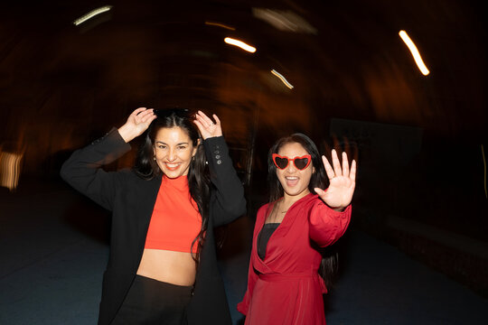 Two smiling women in stylish attire wave and enjoy their walk in the lively ambiance of a city evening