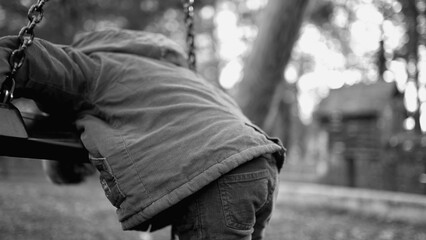 Fototapeta premium Depressed lonely child leaning on park swing feeling sad and gloomy looking down outdoors. One small boy struggling with mental illness in monochromatic, black and white