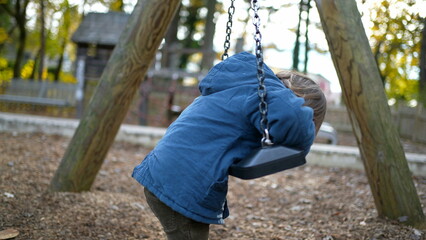 Carefree little boy leaning on park swing during autumn day wearing blue jacket, child twisting and turning into swing playing by himself during fall season outdoors