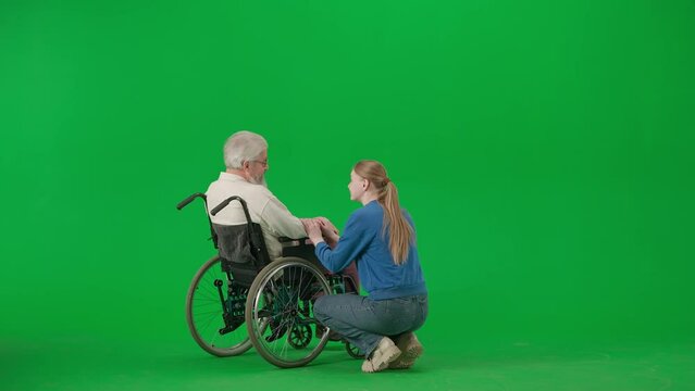 Portrait of aged disabled man on chroma key green screen. Senior man in wheelchair talking with girl assistant or granddaughter.