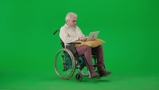 Portrait of aged disabled man on chroma key green screen. Senior man sitting in wheelchair with laptop on the pillow, working.