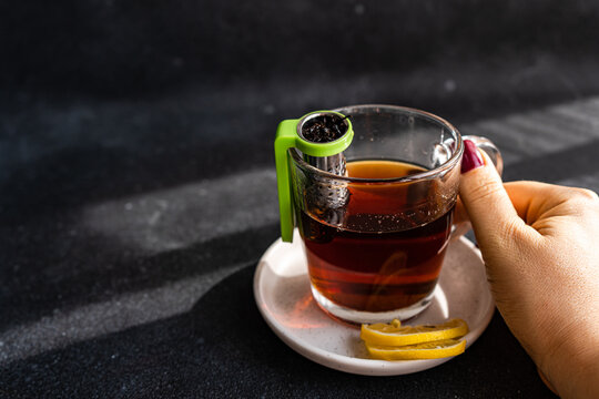 Anonymous hand holding a clear glass cup of tea with a green infuser and lemon slices on a saucer, against a dark background